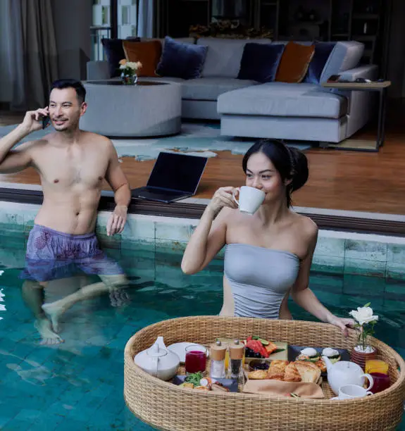 Photo of Happy Asian couple having breakfast in the pool. Floating breakfast tray in luxury hotel. Young woman is drinking morning coffee while her husband talking on the phone, wearing swimsuits