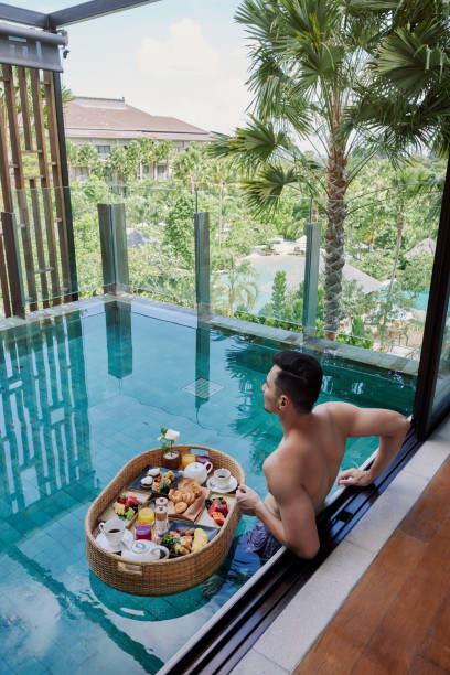 happy man in swimsuit having floating breakfast tray in luxury pool hotel. young male enjoying morning coffee in tropical resort. relaxing, exotic summer travel, holiday, vacation and weekend - bali male beautiful ethnicity imagens e fotografias de stock