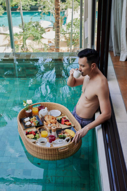 happy man in swimsuit having floating breakfast tray in luxury pool hotel. young male enjoying morning coffee in tropical resort. relaxing, exotic summer travel, holiday, vacation and weekend - bali male beautiful ethnicity imagens e fotografias de stock