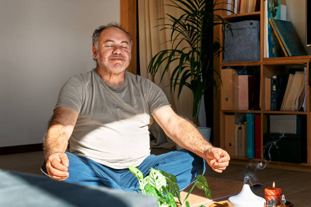 hombre con sobrepeso practicando yoga y meditación en casa sentado en postura de loto en esterilla de yoga, relajado con los ojos cerrados. concepto de meditación consciente. bienestar. - brain gym fotografías e imágenes de stock