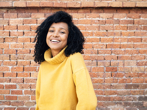 An African woman from Spain in yellow sweater smiling with brick wall on background