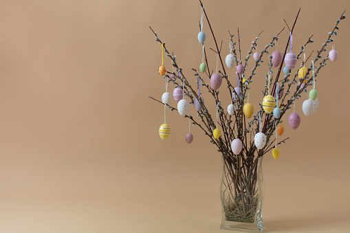 Colored Easter eggs on pussy willow bouquet in glass vase. Front view on beige background with copy space