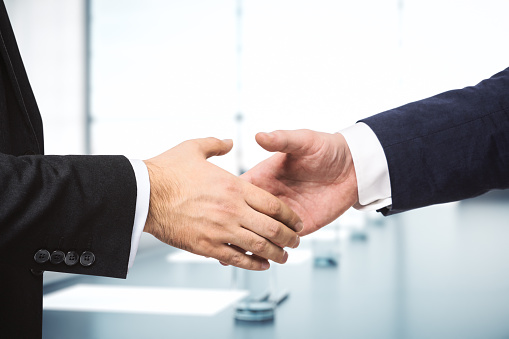 Two businessmen shake hands on the background of sunny meeting room, deal concept, close up