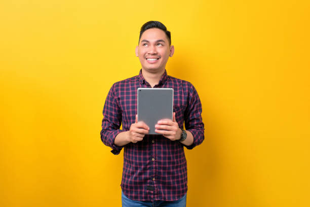 istock Smiling young Asian man holding digital tablet and looking aside at copy space isolated over yellow background 1453946903