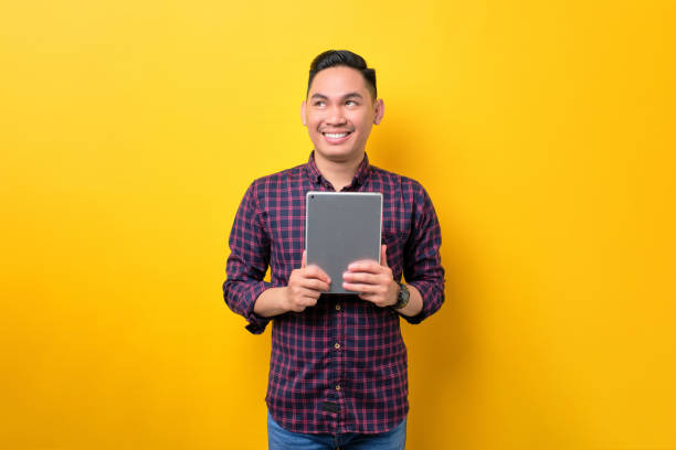 istock Smiling young Asian man holding digital tablet and looking aside at copy space isolated over yellow background 1453946905