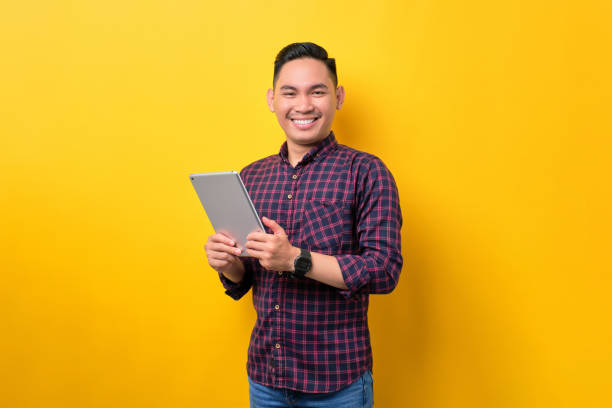 istock Smiling young Asian man holding digital tablet and looking at camera isolated over yellow background 1453946917