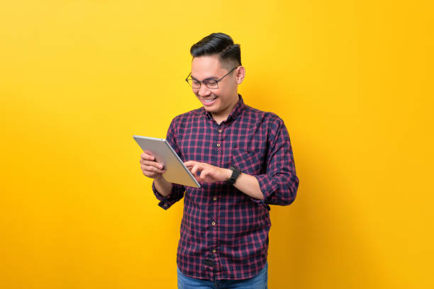 istock Smiling young Asian man in glasses using digital tablet, browsing internet on modern gadget isolated over yellow background 1453946942