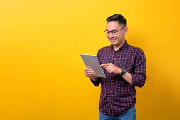 istock Smiling young Asian man in glasses using digital tablet, browsing internet on modern gadget isolated over yellow background 1453946969
