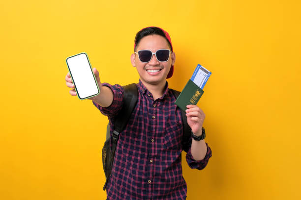 istock Cheerful young Asian man with backpack showing mobile phone blank screen, holding passport and flight tickets isolated over yellow background. Tourism and holiday trip concept 1453947008