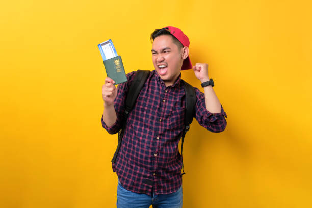istock Excited young Asian man with backpack holding passport and flight tickets, celebrating about trip isolated over yellow background. Tourism and holiday trip concept 1453947020