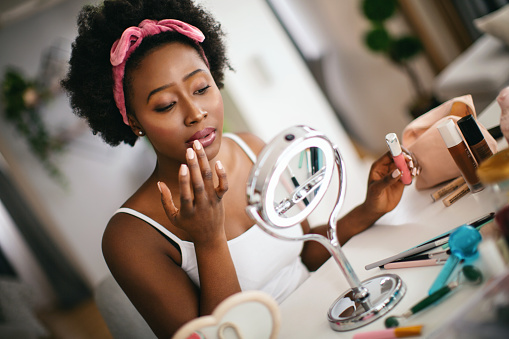 Young woman applying lip gloss with her fingers on her lips while sitting in front of the mirror in her home.