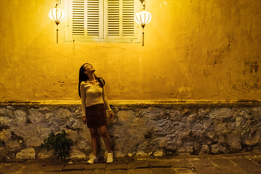 low angle view on young vietnamese woman standing on street in front of illuminated yellow wall in old town of Hoi An and looking up to shining lantern