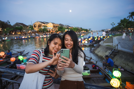 two young woman standing on bridge in old town of Hoi An and looking together at smartphone at sunset hour