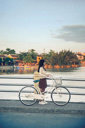side view on young vietnamese woman walking on bridge with bicycle  in old town of Hoi An at sunset hour