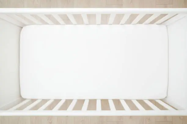 Empty baby crib with white mattress on wooden home floor. Closeup. Top down view.