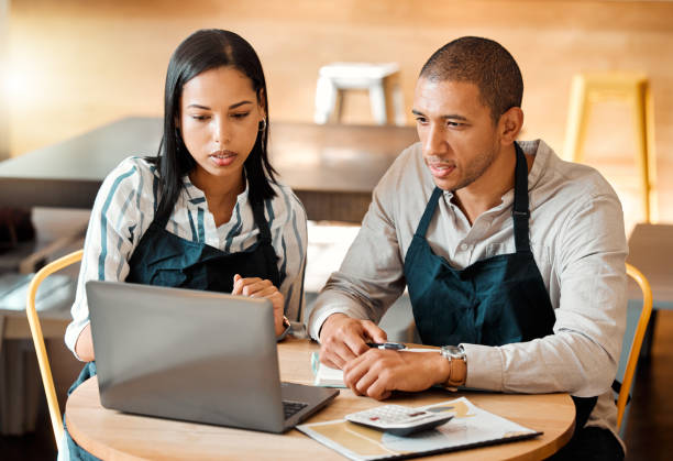 istock Two young mixed race employees wearing aprons sitting at a table in a coffee shop using a laptop and calculator at work. Young male and female hispanic colleagues brainstorming as a team because alone we can do so little, together we can achieve so much 1453950938
