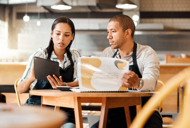 istock Two restaurant workers planning on a digital tablet in a small business. Young mixed race colleagues reviewing plans and reports in a cafe. Colleagues doing digital stocktake and orders with suppliers 1453950942