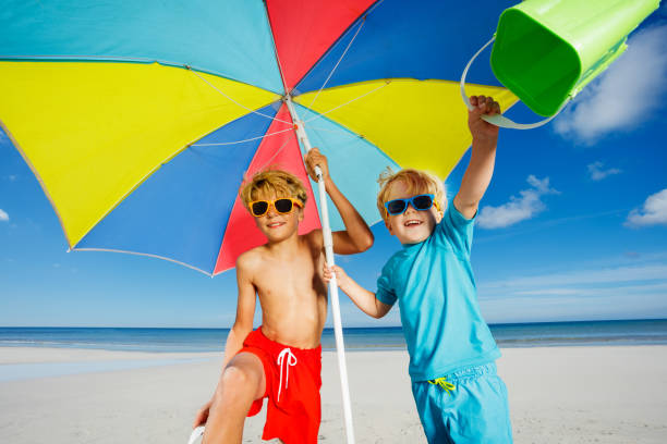 Two cute kids, boys in sunglasses pose on the beach under parasol smiling over ocean on background