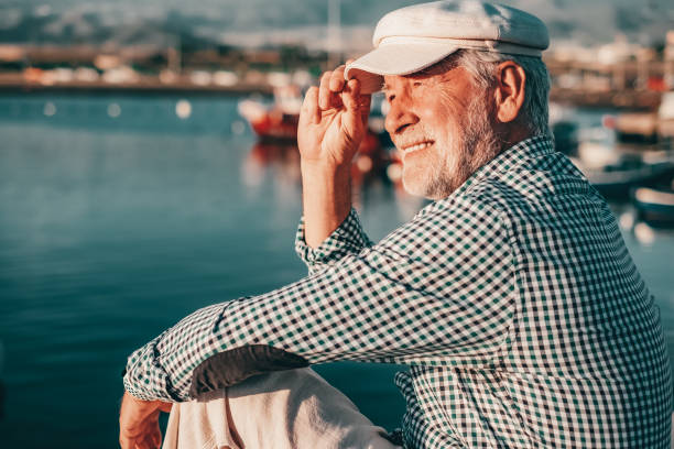 ritratto di uomo anziano felice in camicia casual e cappello seduto al porto di mare al tramonto. nonno barbuto anziano rilassato che si gode il tempo libero, il viaggio o la pensione - fishermen harbor foto e immagini stock