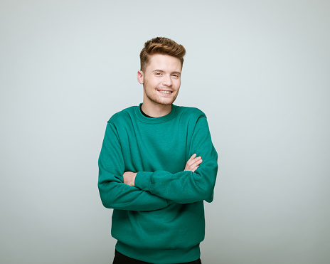 Young man wearing green blouse standing with arms crossed and smiling at camera. Studio shot against grey background.