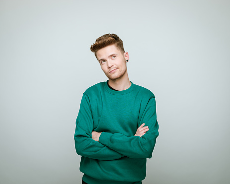 Young man wearing green blouse standing with arms crossed and looking up. Studio shot against grey background.