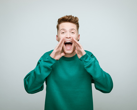 Portrait of young man wearing green blouse shouting at camera with raised hands. Studio shot against grey background.