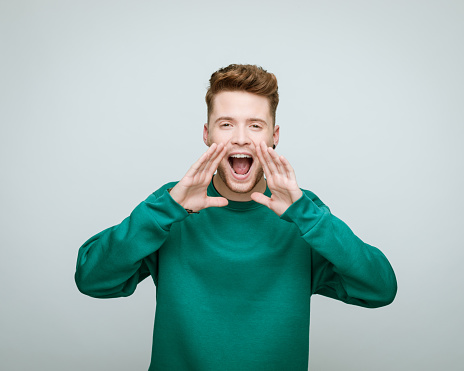 Portrait of young man wearing green blouse shouting at camera with raised hands. Studio shot against grey background.