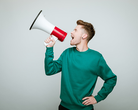 Portrait of young man wearing green blouse shouting into megaphone. Studio shot against grey background.
