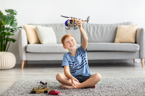 Child redhead boy playing with toys alone at home. Kids play concept. Cheerful boy sitting on carpet in cozy living room, playing with helicopter and cars, enjoying his brand new toys, copy space
