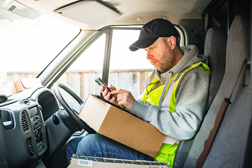 Portrait of a male delivery driver at work. He is sitting in his van looking at a clipboard with documents attached to it and also looking at the screen of a cell phone, with a pile of parcels on the seat and on the dashboard. The man is in his 30s, wearing a high visibility vest, hoodie and baseball cap.