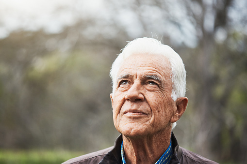 Portrait of senior man in his late 70s, looking upward as he stands outdoors in nature.
