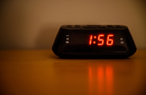 Digital radio alarm clock sitting on a table in a bedroom at night. Dim lighting with shallow depth of field. No logo or branding.
