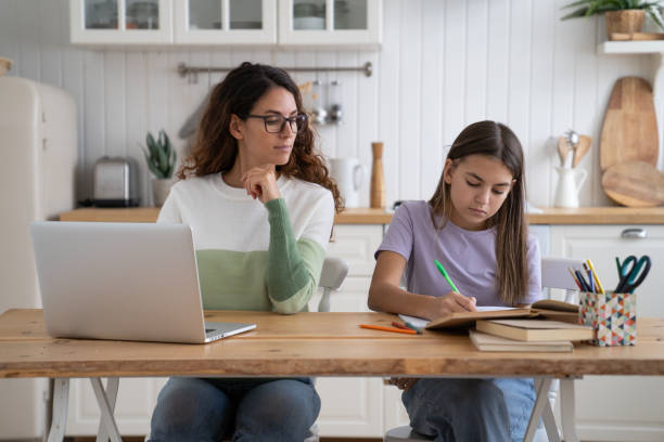 mãe olhando para a filha fazendo a lição de casa enquanto trabalha remotamente ao lado, sentada junta à mesa - homework child mother parent - fotografias e filmes do acervo