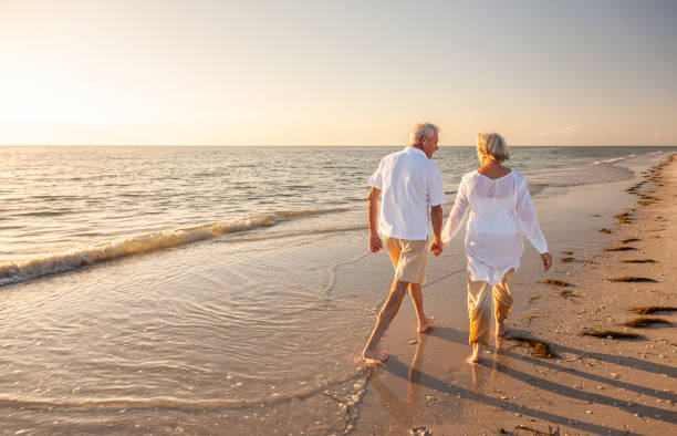 feliz pareja de ancianos jubilados caminando tomados de la mano en la playa al atardecer - senior couple fotos fotografías e imágenes de stock