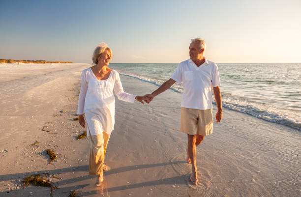 행복한 노인 은퇴한 부부는 해질녘 해변에서 손을 잡고 걷고 있다 스톡 사진