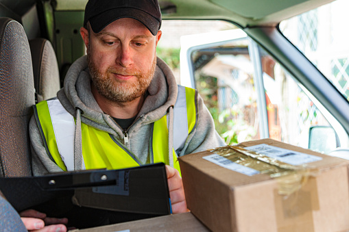 Portrait of a male delivery driver at work. He is sitting in his van with a pile of parcels on the seat and on the dashboard. The man is in his 30s, wearing a high visibility vest, hoodie and baseball cap.