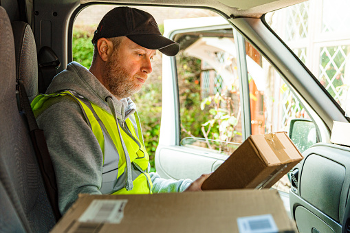 Portrait of a male delivery driver at work. He is sitting in his van with a pile of parcels on the seat and on the dashboard. The man is in his 30s, wearing a high visibility vest, hoodie and baseball cap.