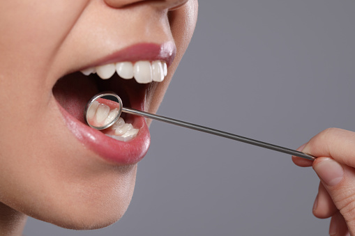 Dentist examining woman's teeth with mirror on grey background, closeup