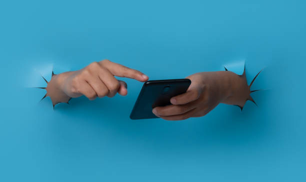 istock Female hand holding smartphone and touching screen breaks through blue paper background. 1453985711