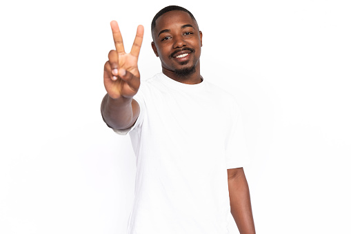 Happy African American man showing victory sign. Portrait of pleased young male model with short hair in white T-shirt looking at camera, smiling, greeting someone. Success, optimism concept
