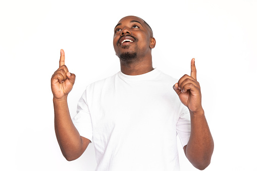 Happy African American man pointing up. Portrait of pleased young male model with short hair and beard in white T-shirt looking up, showing ad with fingers, smiling. Advertising concept