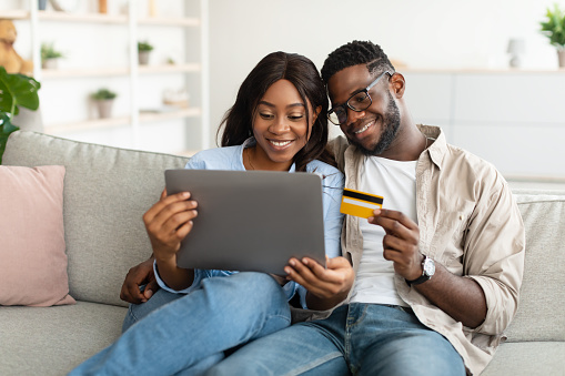Online Shopping Concept. Portrait of millennial African American couple sitting on couch at home and choosing what to buy, excited woman holding digital tablet, guy in glasses using credit card