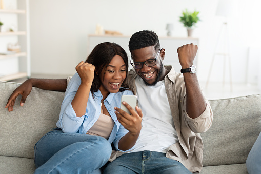 Portrait of excited African American couple using smartphone, young lady and guy making winner sign gesture, shaking fists, sitting on couch in living room, playing video games or reading great news