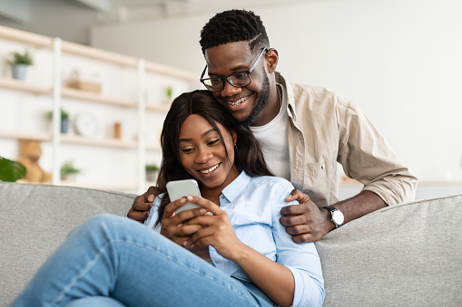 Smiling young black man in braces embracing woman from behind while looking at smartphone. African American couple sharing social media on cellphone, happy female sitting on the sofa, showing gadget