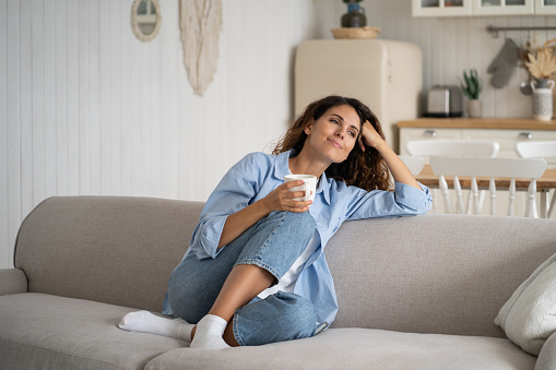 Young happy relaxed Latin woman enjoying quiet moments at home with cup of tea or coffee, smiling female holding mug with hot drink resting on sofa in living room and dreaming, relaxing of everything