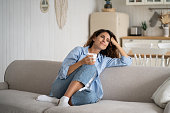 Young happy relaxed woman sitting on sofa enjoying quiet moments at home with cup of tea or coffee
