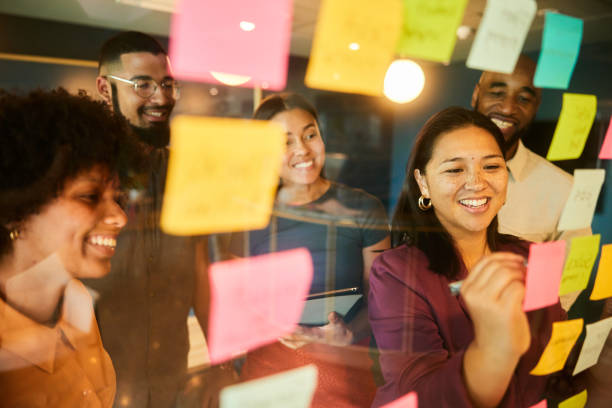 Businesspeople laughing while brainstorming with adhesive notes on an office window Diverse group of businesspeople laughing during a brainstorming session using adhesive notes on a glass wall in an office brainstorming stock pictures, royalty-free photos & images