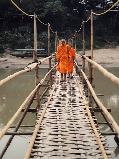 Luang Prabang, Laos in March 2019: Two Buddhist monks crossing Nam Khan River in Luang Prabang on a bamboo bridge.\nShot on iPhone 6, Processed with VSCO with s2 preset