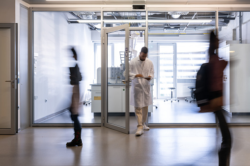 A chemical worker in a white coat stands between the doors of the faculty laboratory, students rush past him in the corridor. Blurred people while moving on the photo.