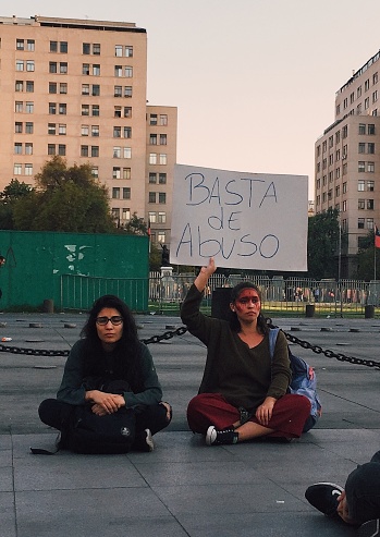 Santiago de Chile, October 2019: Two female protestors sitting in front of La Moneda, the Chilean president's residence. One of them is holding up a sign saying 'Basta de abuso', translating to 'enough with the abuse'.